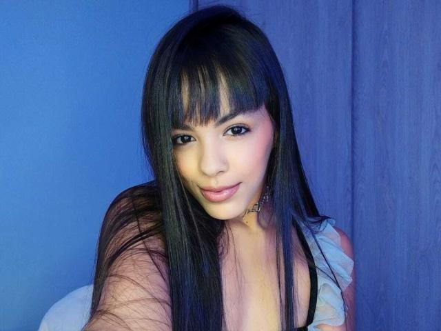 MagiicBebes a de longs cheveux noirs avec une frange, des yeux foncés et du rouge à lèvres rose, et elle sourit légèrement. Elle porte un haut noir avec une épaule froncée, qui révèle ses petites seins..