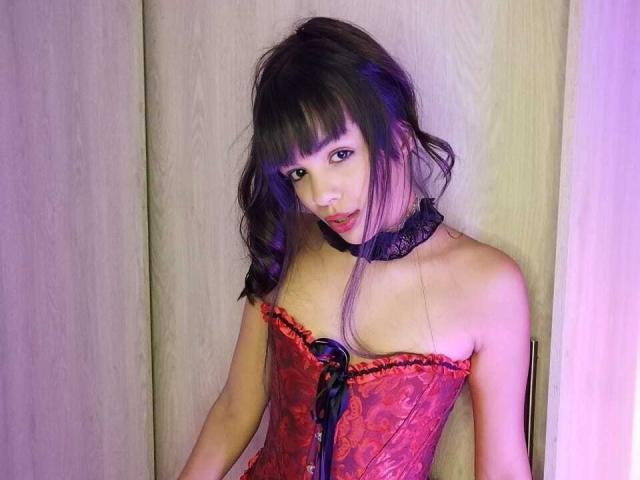 MagiicBebes avec des cheveux foncés et une frange, portant un corset en dentelle rouge avec un ruban noir, un collier noir et des mèches violettes, regardant séductriceusement l'appareil photo..