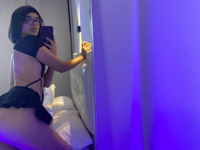 MagiicBebes, vêtue de lingerie noire avec une bretelle sur le côté, a une jolie fesse, et se regarde dans un miroir, tenant un téléphone, dans une chambre éclairée en violet..