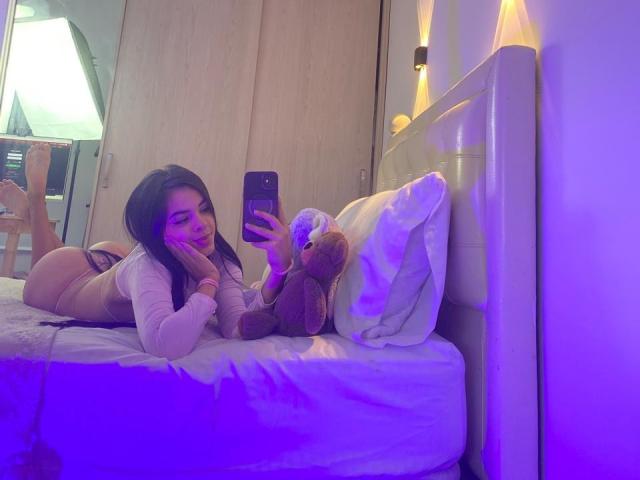 MagiicBebes lag auf einem Bett, machte ein Selfie mit einem Handy, trug ein weißes Oberteil mit langen Ärmeln und zeigte ihren Hintern. Neben ihr lag ein brauner Teddybär..