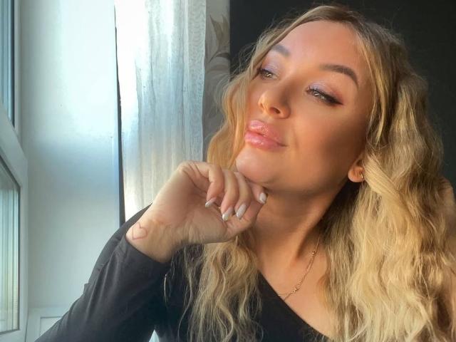 DemiMoveX with pouty lips, blonde wavy hair, subtle pink eyeshadow, and glossy pink lipstick, wearing black top, hand on cheek, sunlight on left, calm expression.
