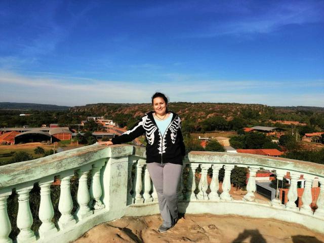 LadySelene with medium tits, black and white patterned jacket, gray pants, standing on a green, weathered balcony, smiling, arms out, blue sky, and distant green hills in background.