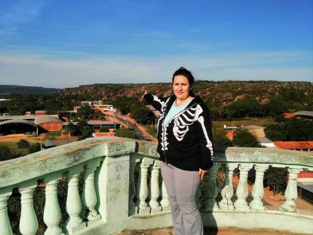 LadySelene stands on a green, weathered balcony in a black skeleton hoodie, gray pants, and white shirt, with a clear blue sky and green hills in the background.