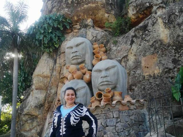 LadySelene stands in front of stone carvings with large faces, surrounded by clay pots. She's wearing a black skeleton hoodie, smiling, with her hands on her hips.