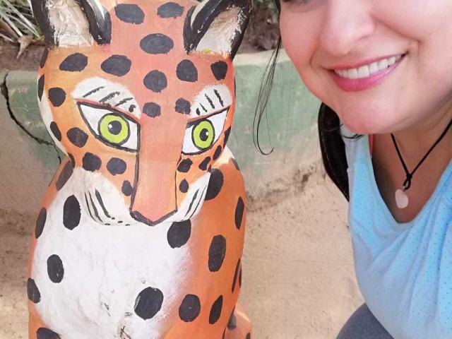 LadySelene's face is next to a painted, spotted, cat-like statue with green eyes. She's smiling with pink lipstick, wearing a blue shirt and a necklace.