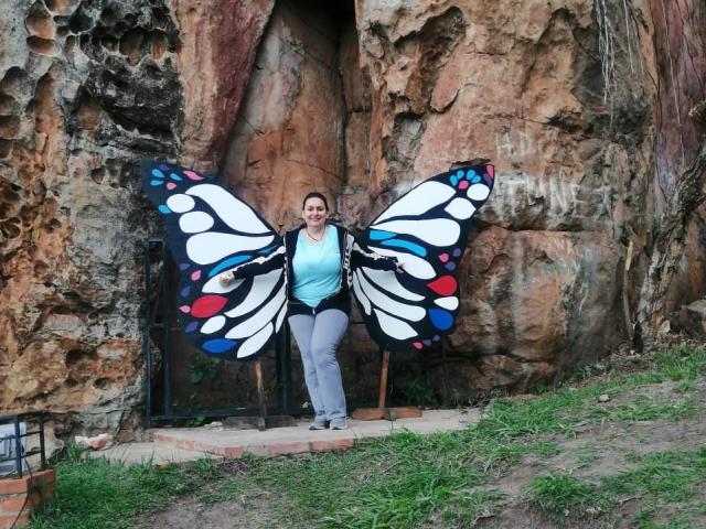 LadySelene with large, colorful butterfly wings standing in front of a rocky wall, wearing a blue top and gray pants, smiling with brown hair tied back.