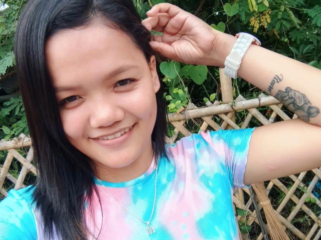 SexyCleopatraForU with black hair, smiling, wearing a pink-blue tie-dye shirt, white wristwatch, and visible tattoo on her right forearm, holding her hair up.
