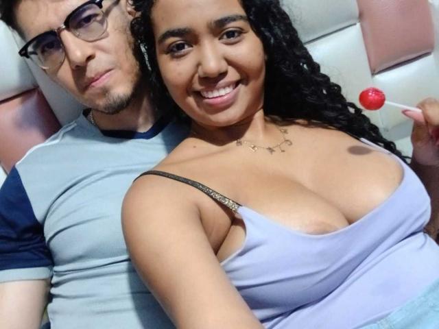 LeoAndKali with curly black hair, tan skin, and big tits in a purple top, holding a red lollipop, smiling. LeoAndKali with glasses, blue and gray shirt, behind her.
