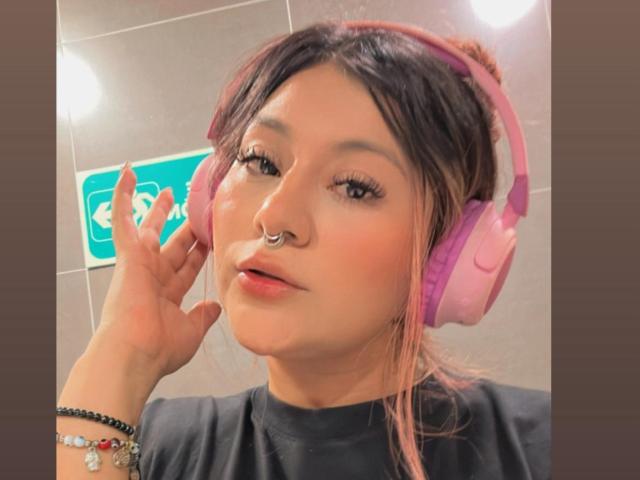LorraineV with pink headphones, nose ring, and black shirt, looking at camera with pink hair, subtle makeup, and black eyeliner, showing off her manicured fingers.