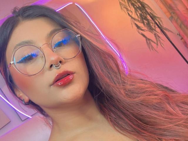 LorraineV with round glasses, blue eyeliner, nose ring, red lips, glossy, and long brown hair, looking at camera, neon pink and purple light in background.