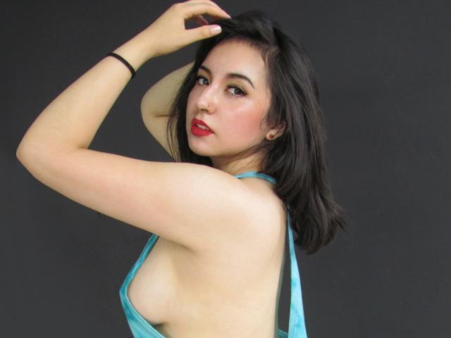 SusanMonroy with black hair, red lipstick, and a blue halter top, exposing her titty, raising her arm, and looking at the camera. Black background.