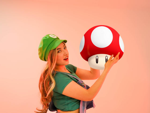 SusanMonroy with long blonde hair, green hat, green shirt, and purple overalls, holding a giant red mushroom with white spots, smiling, pink background, looking at the mushroom.