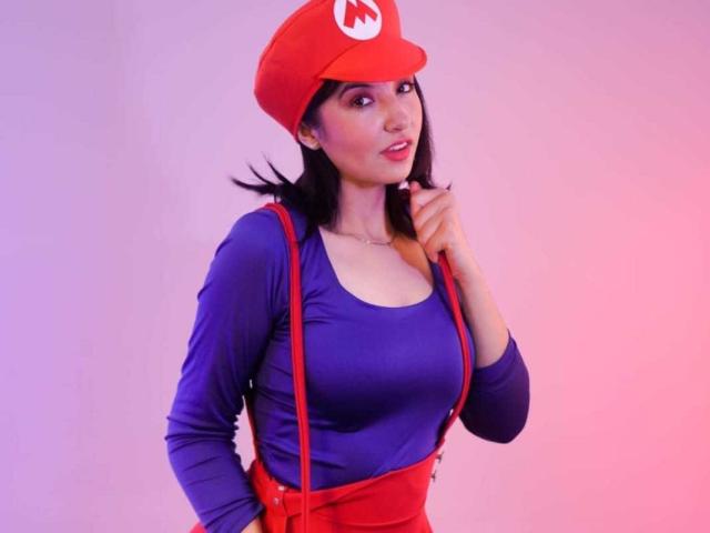 SusanMonroy with dark hair, red Mario hat, blue long-sleeve shirt, red suspenders, and red pants, touching her chin, pink and purple lighting, showing her tits and ass.