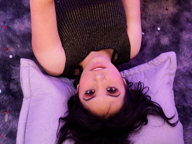 SusanMonroy is lying on her back with her head upside down, wearing a sparkly black sleeveless top, and looking up at the camera with a cute smile.