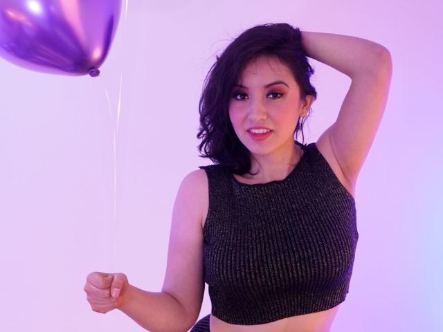 Susan Monroy, con i capelli scuri e ondulati, indossa una crop top nera scintillante, tiene in mano un palloncino viola, sorride, con il braccio destro alzato e il braccio sinistro che stringe il filo del palloncino..