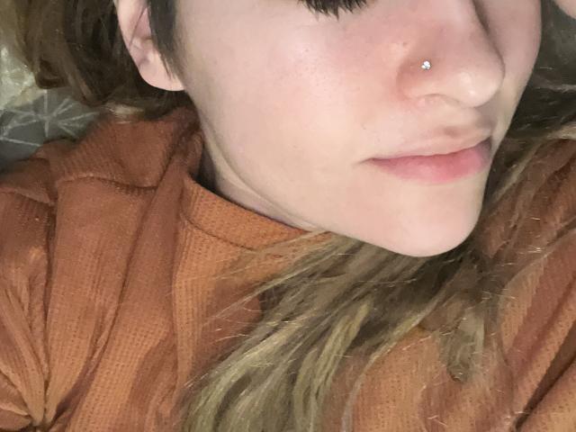 ArgentineHotCouple's nose has a small silver piercing, with light pink lips slightly parted, wearing an orange textured shirt, and blonde hair partially visible.