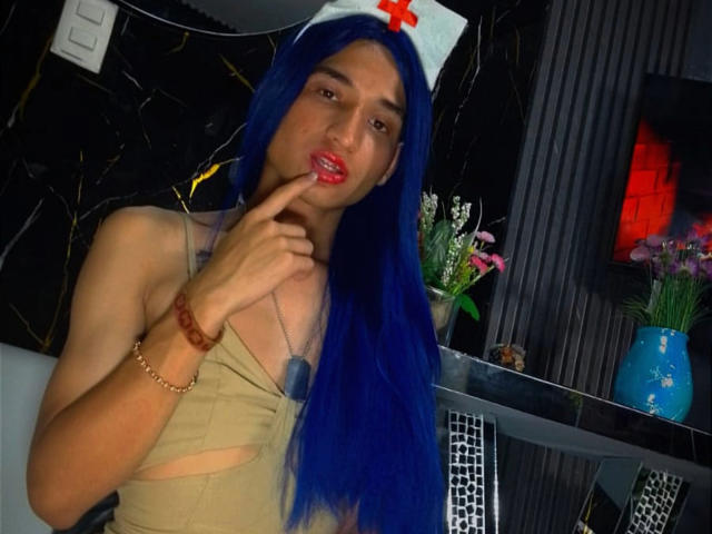 SaraLatin69 with blue wig, red lipstick, and dog tag, sucking finger, in beige top, wearing nurse hat, tan skin, slim build, standing in dark room, holding vase.