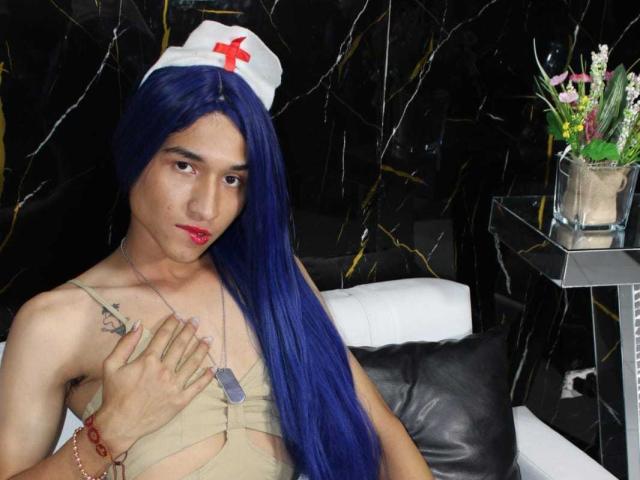 SaraLatin69 with blue wig, nurse hat, tan dress, hand on chest, dog tags, red lipstick, tattoo on left shoulder, black marble wall, white couch, vase with flowers.