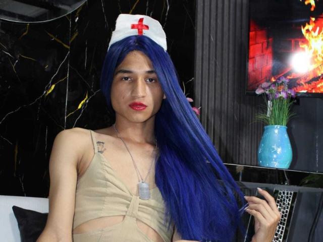 SaraLatin69 with long blue hair, wearing a nurse hat with a red cross, beige top, dog tags, and red lipstick, holding her hair, showing a small tattoo on her chest.