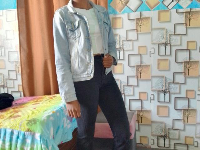 Nerina with a denim jacket, white shirt, black high-waisted jeans, standing, hand on waist, head out of frame, next to a colorful bedspread, patterned wall in background.