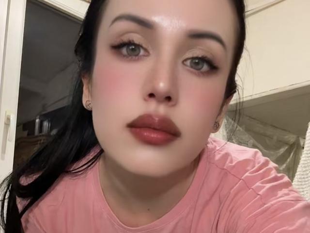 BeautyCoral's face is centered with pinkish cheeks, full red lips, green eyes, black eyebrows, and black hair in a ponytail, wearing a pink shirt, looking at the camera.