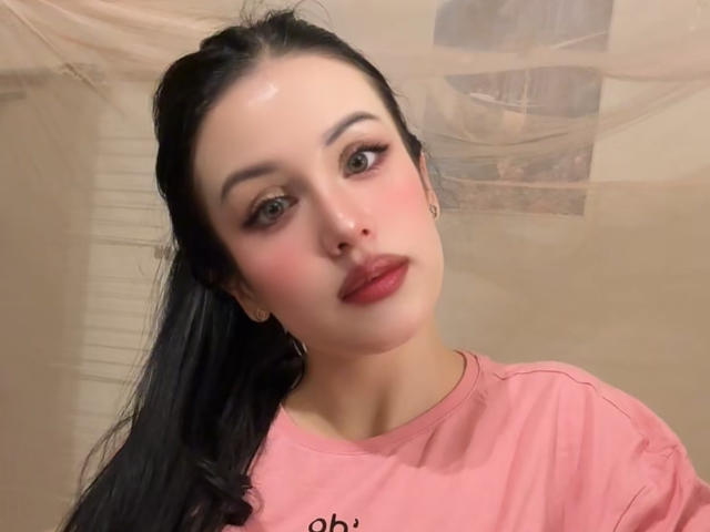 BeautyCoral with shiny skin, black hair, pink shirt, red lipstick, and hoop earrings, looking up with makeup highlighting her eyes. Her tits are not visible.