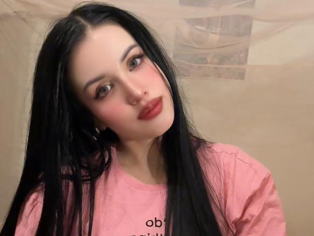 BeautyCoral with long black hair, pink shirt, red lipstick, and hoop earrings, looking slightly to the side with a subtle smile. Soft pink blush on her cheeks.