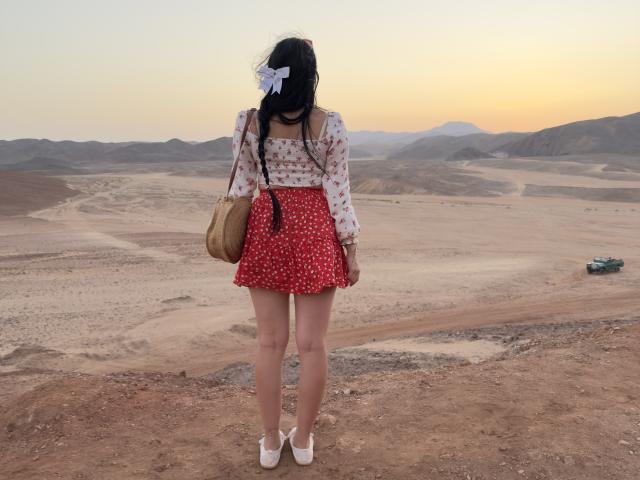 BeautyCoral stands with her back to the camera, wearing a red polka dot skirt, white floral blouse, white shoes, tan bag, and a white bow in her black hair.
