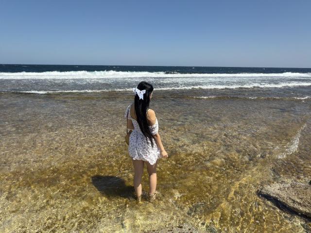 BeautyCoral stands in shallow, clear water, facing the ocean. She's wearing a white, off-shoulder dress with a large bow in her long black hair. Her ass is visible.