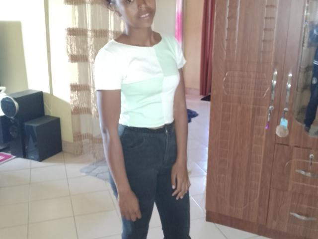 Nerina with dark skin, small tits, white and green striped shirt, black jeans, standing in a room, wooden wardrobe on the right, smiling slightly.