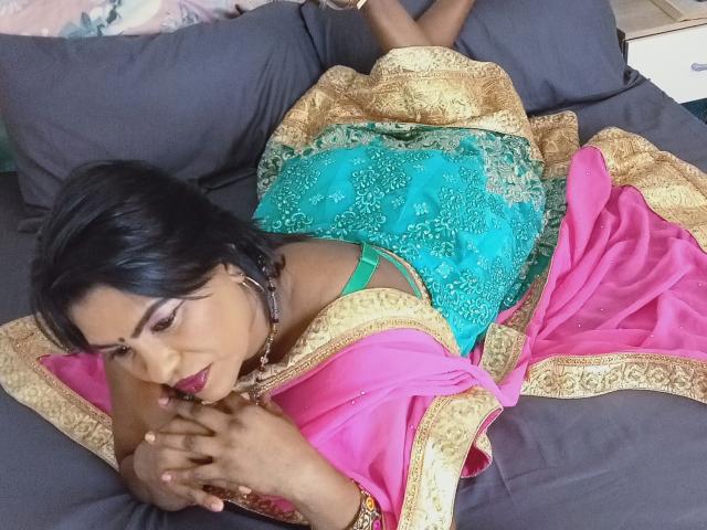 Aadhya est allongée sur un lit, vêtue d'un haut en dentelle bleu turquoise et d'un sari rose orné de détails dorés, laissant entrevoir ses fesses et son soutien-gorge vert, avec des cheveux noirs et du rouge à lèvres..