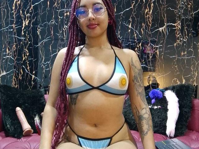 AryVixen, com tranças vermelhas, óculos e tatuagens, vestindo um biquíni azul e branco com estampas de sol amarelas, sentada em um sofá rosa com um brinquedo sexual e um travesseiro preto fofo..