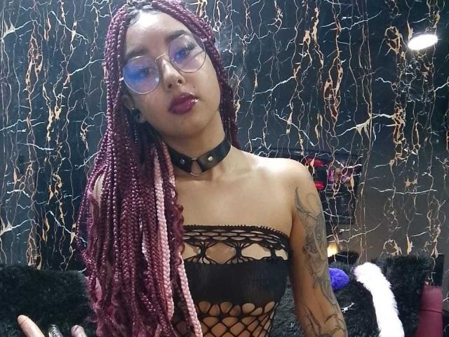 AryVixen with maroon and pink braids, glasses, dark lipstick, black mesh top showing tits, tattooed arm, black choker, and raised eyebrows. Dark, textured background.