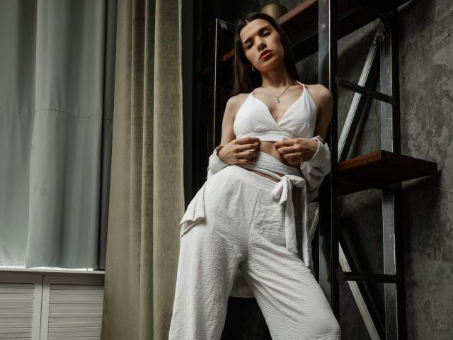 LinnieFlower with small tits, wearing a white crop top and pants, standing against a metal shelf, holding her top, looking down, in a room with curtains.