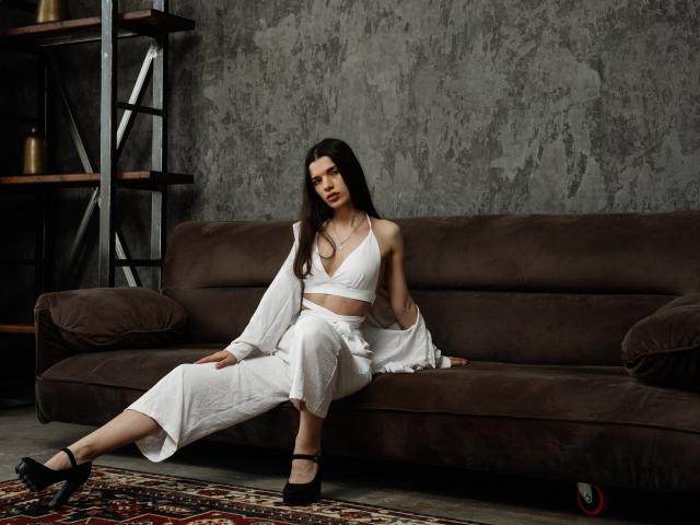 LinnieFlower sitting on a brown sofa, wearing a white halter top and pants, with a loose shirt, black heels, and long dark hair, looking at the camera.