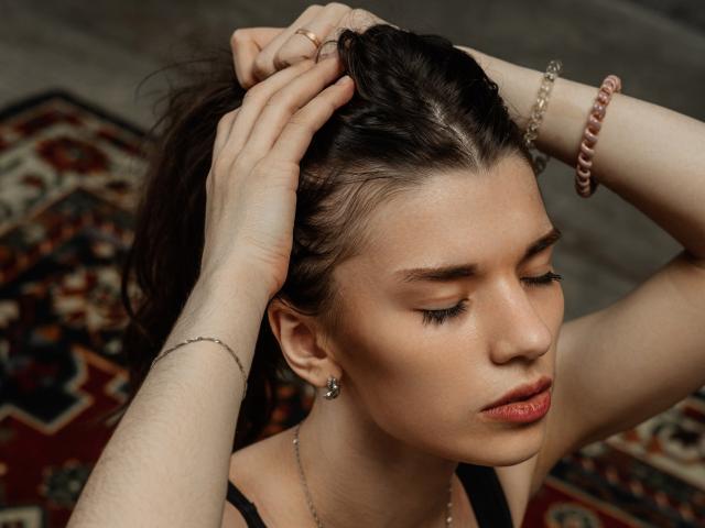 LinnieFlower has her hands in her brown hair, showing off her tits and ass in the background, with eyes closed and red lipstick, wearing a black top and pink bracelet.