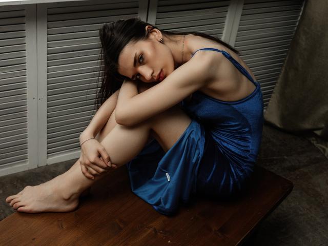 LinnieFlower sits on a wooden table, legs crossed, arms wrapped around knees, wearing a blue, shiny dress, looking down with a slight, melancholic smile. Her tits are small.