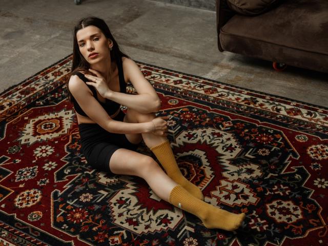 LinnieFlower with small tits, sitting on a red and black patterned rug, wearing a black top and shorts, yellow socks, looking off-camera, in a room with a brown sofa.