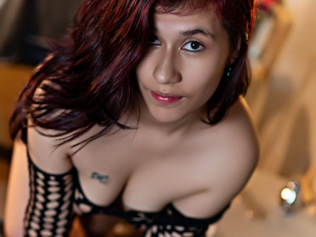 MillaCasanova with red hair, winking, wearing black fishnet top, showing tits, small tattoo on left chest, blue earrings, looking down, soft smile, slightly blurred background.