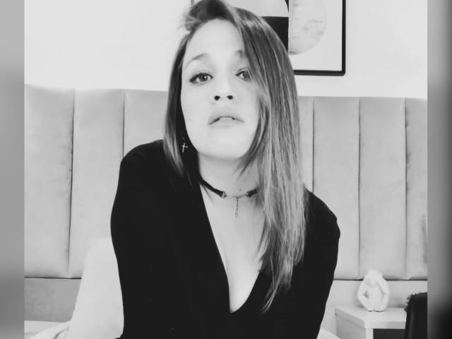 Black and white photo of MillyGrey with straight, shoulder-length hair, wearing a black top and choker, looking up with a small, cute nose and slight smile.