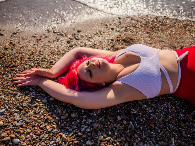 CoolTatis with bright pink hair, lying on a rocky beach, arms above head, wearing a white sports bra, showing her big boobs, and red pants, eyes closed, sunlit.