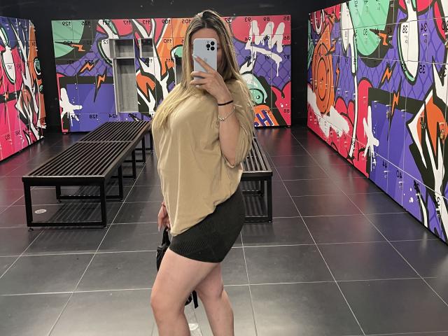JennyLawry, avec de grosses fesses, portant un haut beige et une mini-jupe noire, tenant un téléphone, debout devant des murs recouverts de graffitis colorés, se prenant un selfie dans une pièce carrelée..