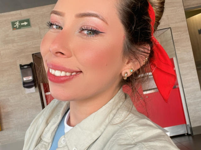 JennyLawry's face with red lipstick, pink eyeshadow, and a red headscarf, smiling with white teeth, wearing a green jacket, and a blue shirt, in a bathroom.