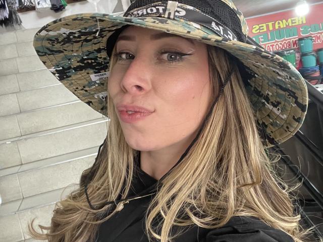 JennyLawry with blonde hair, wearing a camo sunhat, black eyeliner, and black jacket, pouting slightly, looking at camera, in a store.