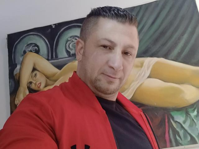 KamJhonson, a chubby guy with short dark hair and stubble, wearing a red jacket over a black shirt, stands in front of a painting of a naked woman with large tits.