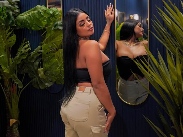 GabrielleKing with long black hair, tan skin, and large ass in a black strapless top and beige pants, touching a mirror, showing her reflection and a confident expression.