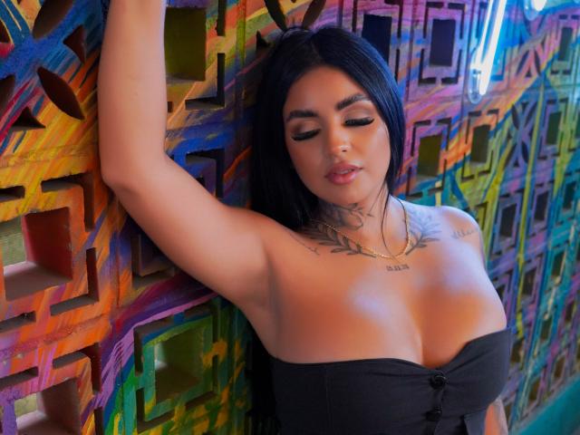 Gabrielle King, con capelli neri, seno abbondante e tatuaggi, indossa un top nero senza spalline, con le braccia sollevate sopra la testa, appoggiata a un muro colorato con motivi geometrici. In secondo piano, una luce al neon..