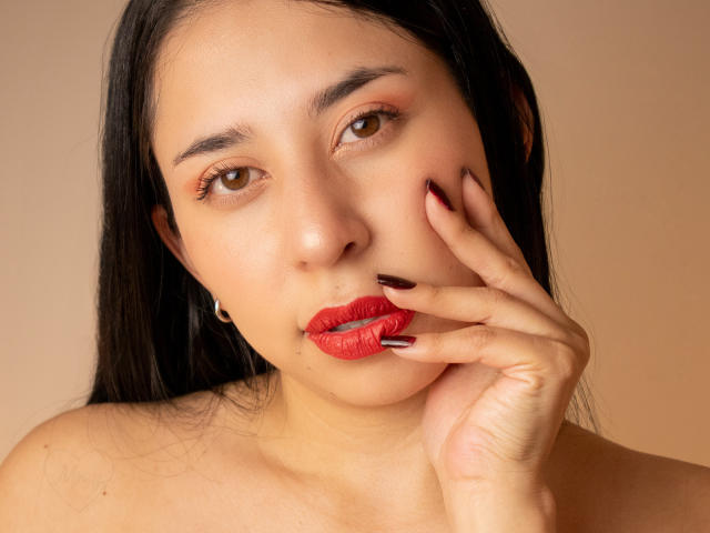 VanesaSwift has red lipstick, brown eyes, black hair, and dark red nails, touching her lip with her hand, wearing a silver hoop earring, against a beige background.