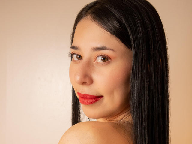 VanesaSwift hat eine glatte, helle Haut, roten Lippenstift und langes, schwarzes Haar. Sie blickt mit braunen Augen und einem leichten Lächeln zurück und zeigt ihre nackten Schultern..
