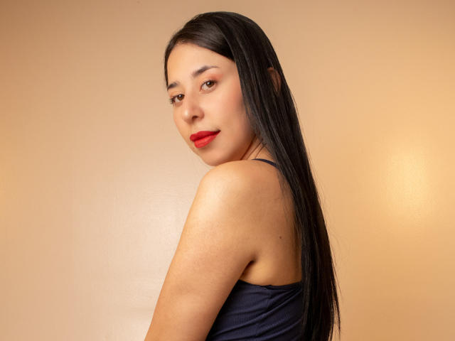 VanesaSwift tiene el pelo largo y negro, lápiz labial rojo y una sonrisa encantadora. Lleva una blusa azul oscuro que deja al descubierto su hombro. Está mirando por encima del hombro, con una expresión sensual..